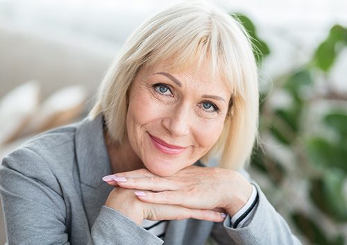 Lovely middle-aged blond woman with beaming smile sitting on sofa and looking at camera